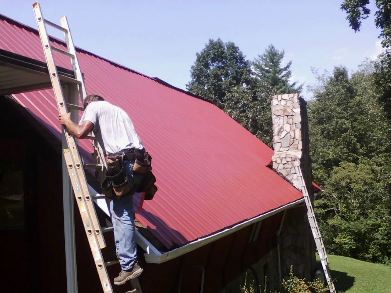 Roofer on a ladder beside a red metal roof and stone chimney — Soffit & Fascia Repair by Roofing Brothers in Meadow